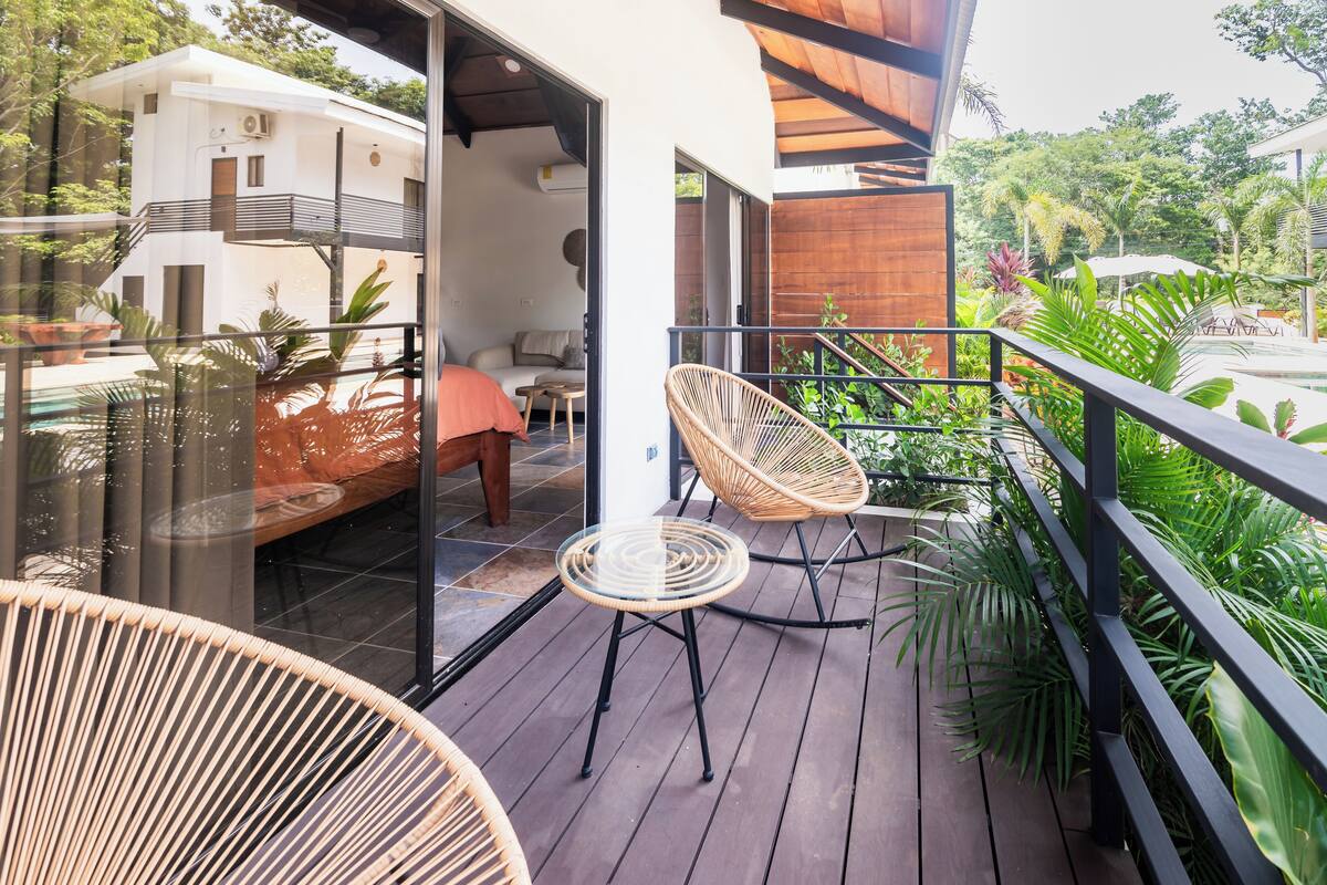 Una terraza de madera con sillas de mimbre, una mesa de vidrio y vista a un edificio blanco con balcón.
