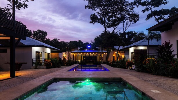 Un área de piscina moderna con una piscina cristalina, rodeada de jardines bien cuidados y un edificio con entrada iluminada en azul.