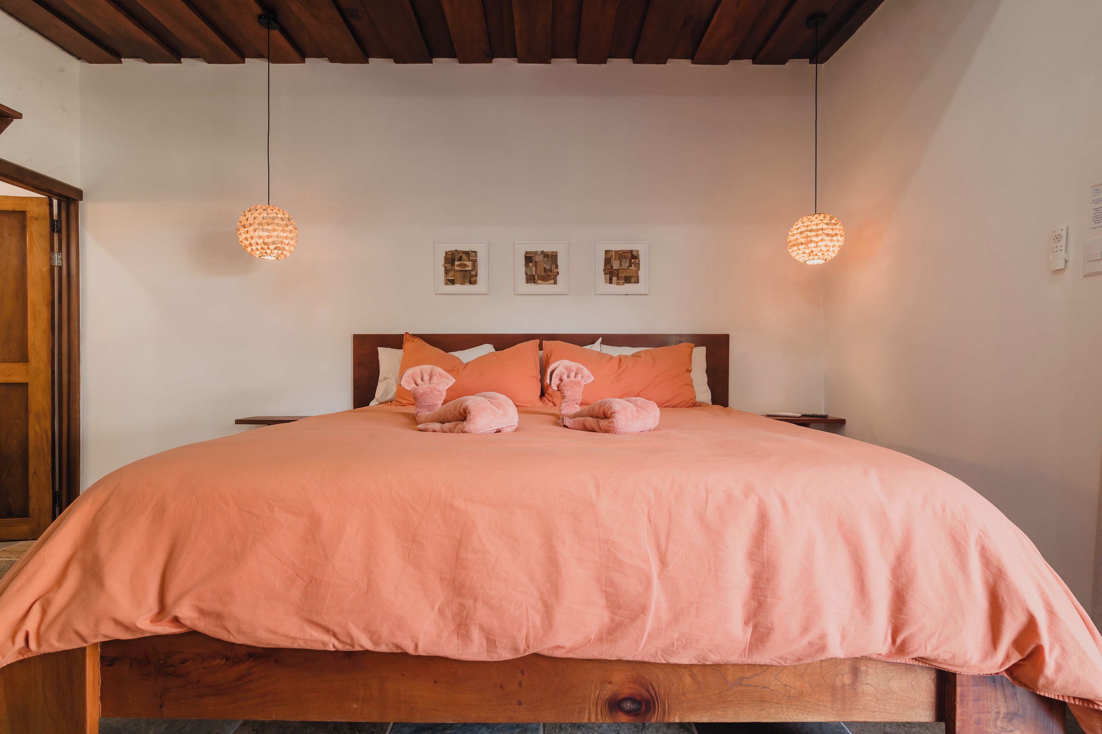Una cama bien hecha con colcha y almohadas rosa, dos lámparas colgantes y tres cuadros en la pared.