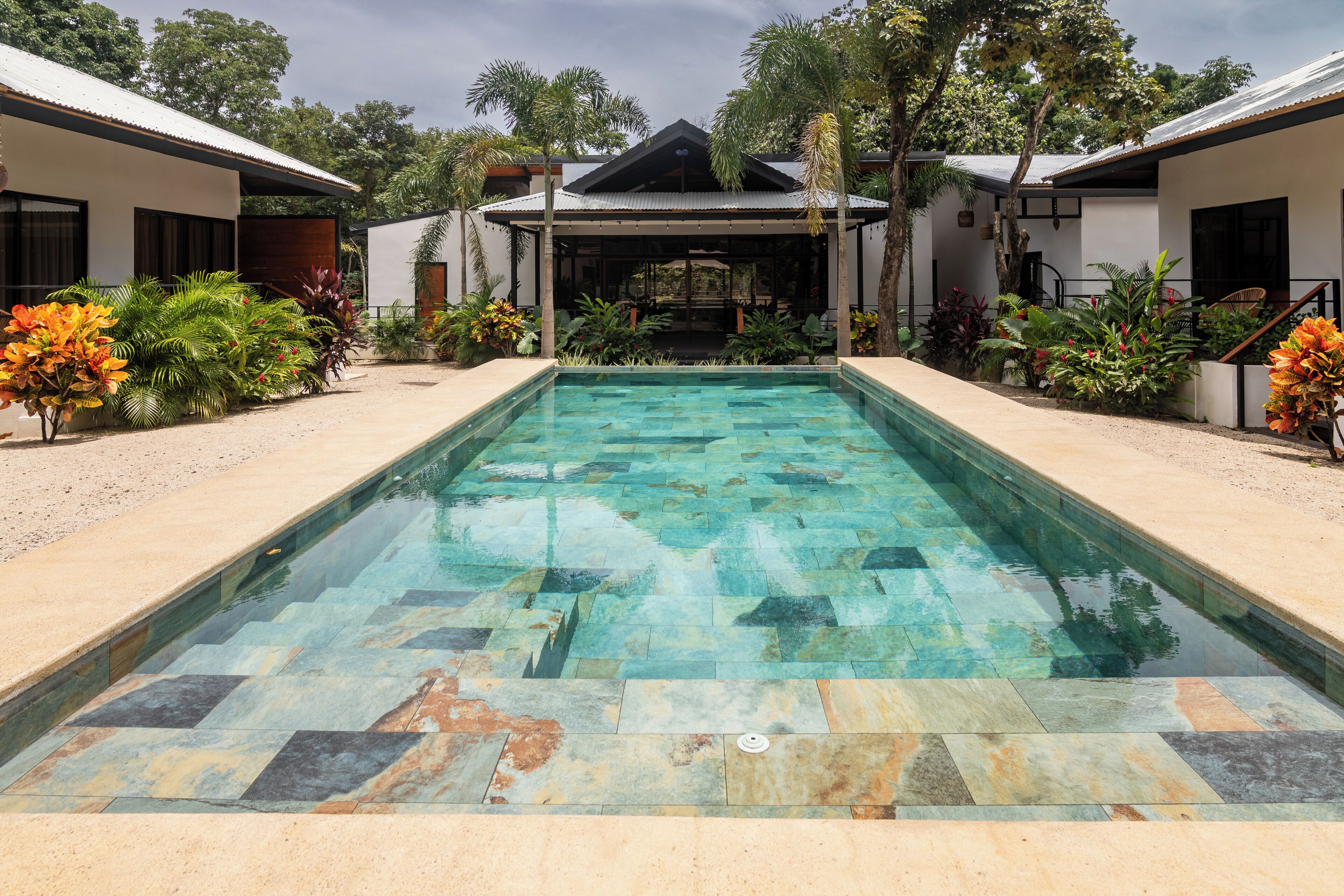Une piscine rectangulaire bordée de carreaux de pierre, entourée d’une zone sablonneuse et de plantes tropicales.