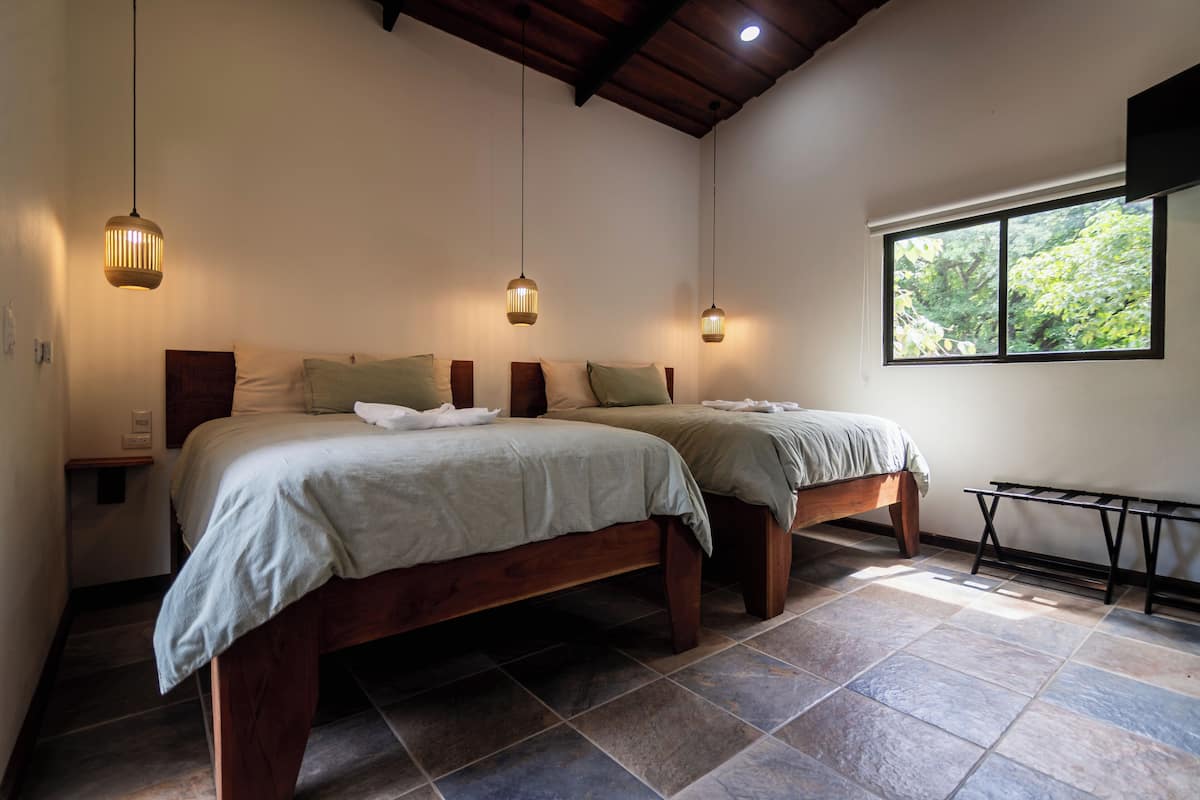 Un dormitorio con dos camas de madera, una ventana con vista a árboles y lámparas colgantes.