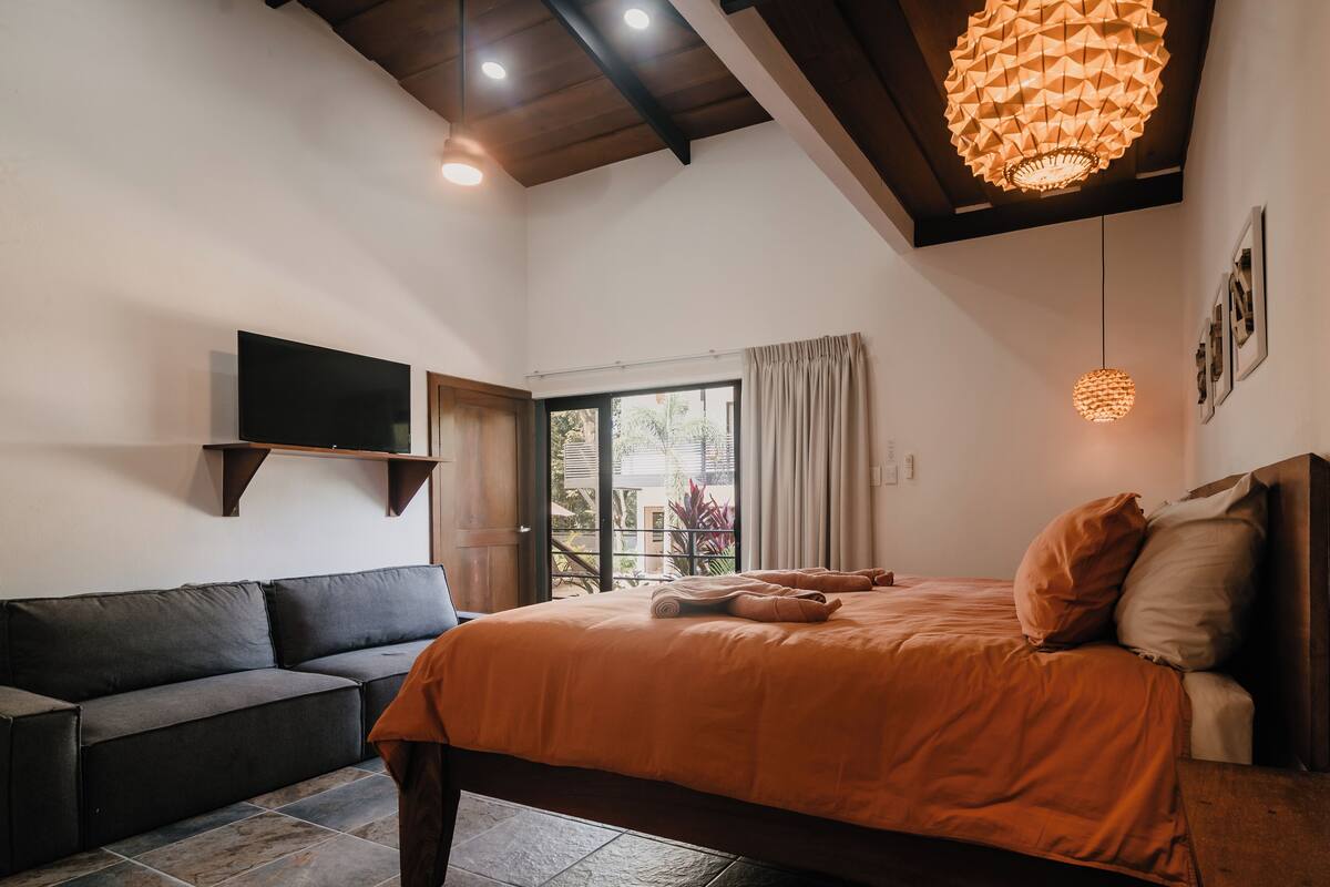 Un dormitorio con una cama, un sofá, un televisor y un ventanal con cortinas.