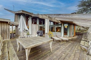 Een rustiek houten terras met een tafel en stoelen, omringd door gebouwen met hout en grote ramen en een overdekte zithoek.