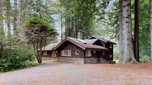 Une cabane rustique, dotée de fondations en pierre et d’un revêtement extérieur en bois, entourée de grands arbres et accessible par une allée de gravier.