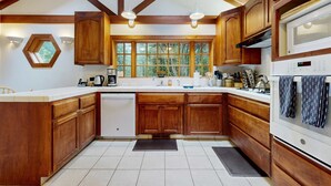 Une cuisine avec des meubles en bois, un plan de travail blanc, un lave-vaisselle encastré et une fenêtre donnant sur des arbres.