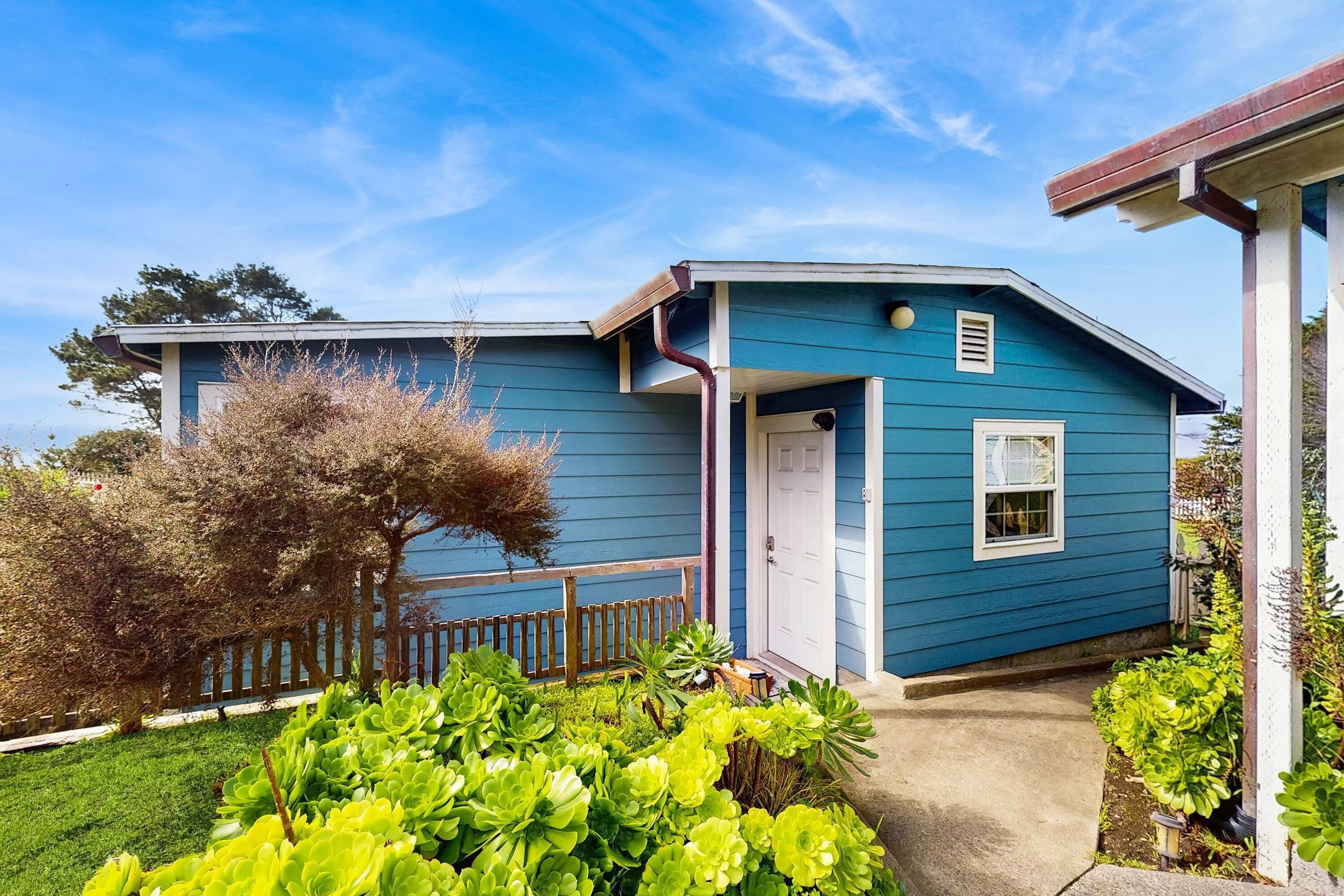 Une maison bleue avec un porche à l’avant, une clôture en bois et des plantes vertes dans le jardin.