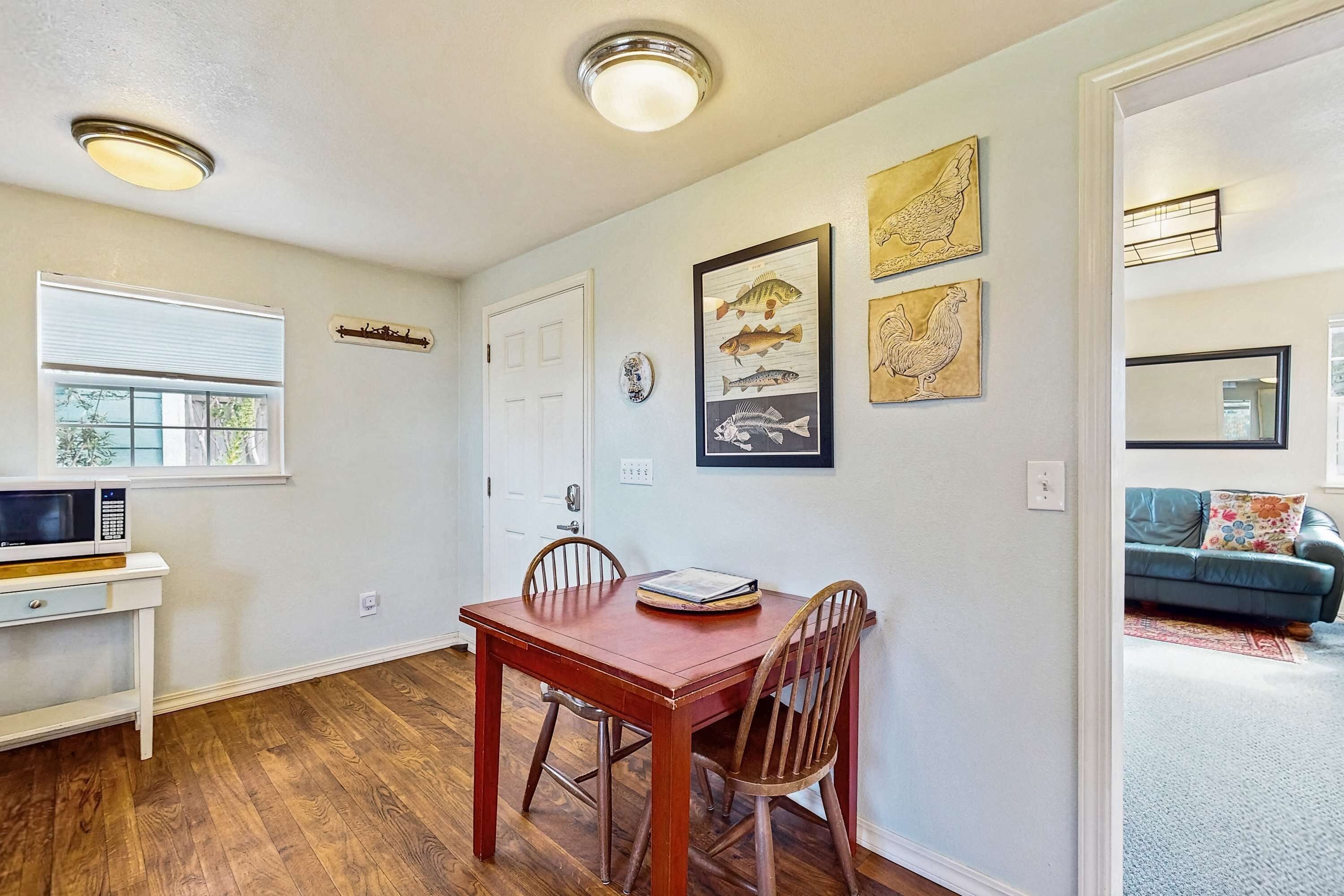 Un comedor con mesa y sillas de madera, pared blanca con obras de arte enmarcadas, ventana con persianas y un microondas sobre un mueble blanco.
