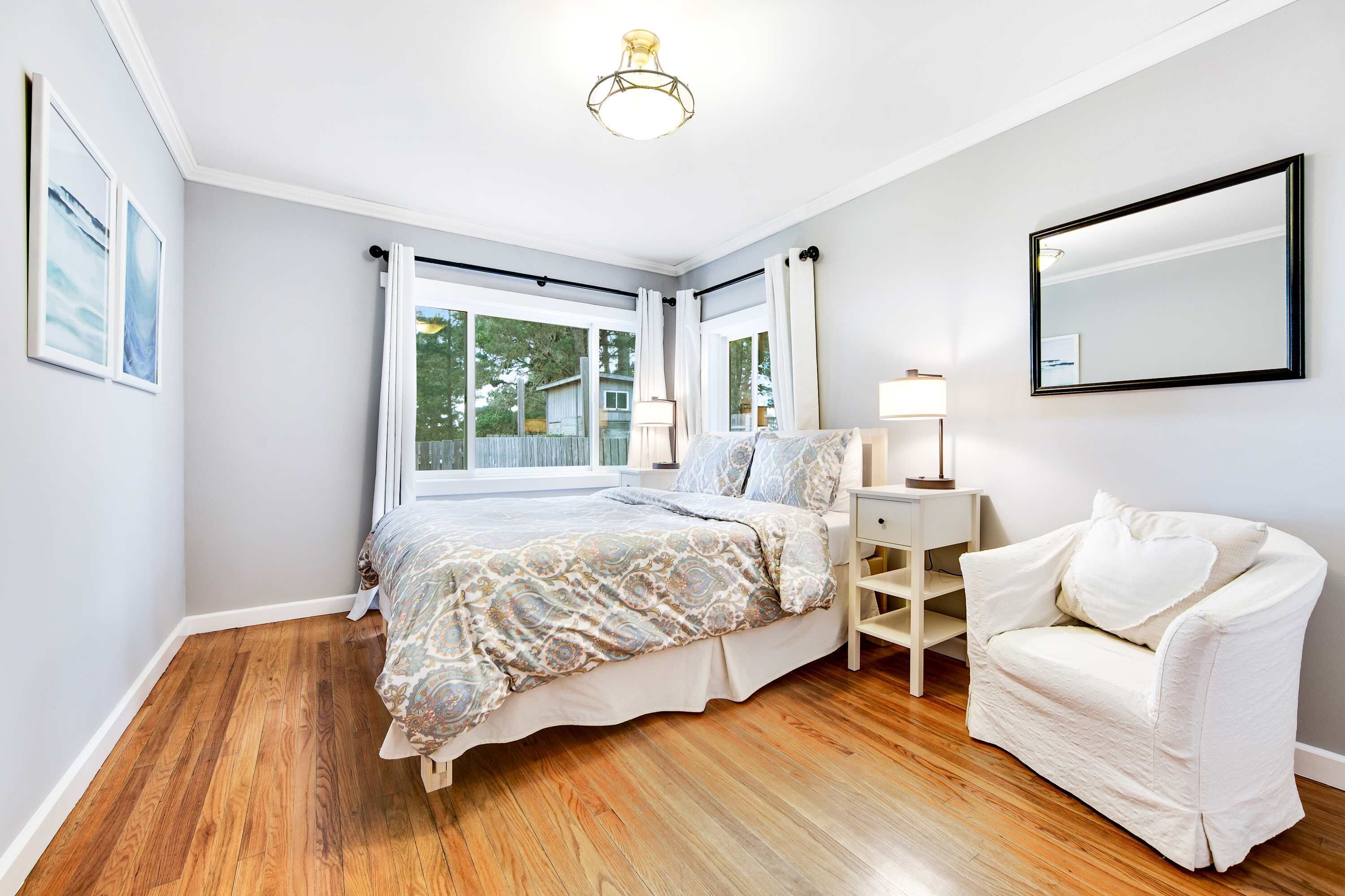 A bedroom with a bed, a white armchair, a bedside table with a lamp, and a large window with curtains.