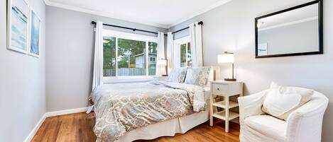 Ein Schlafzimmer mit einem Bett, einem weißen Sessel, einem Nachttisch mit Lampe und einem großen Fenster mit Vorhängen.