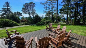 A wooden deck with Adirondack chairs and a table, surrounded by lush greenery and tall trees.