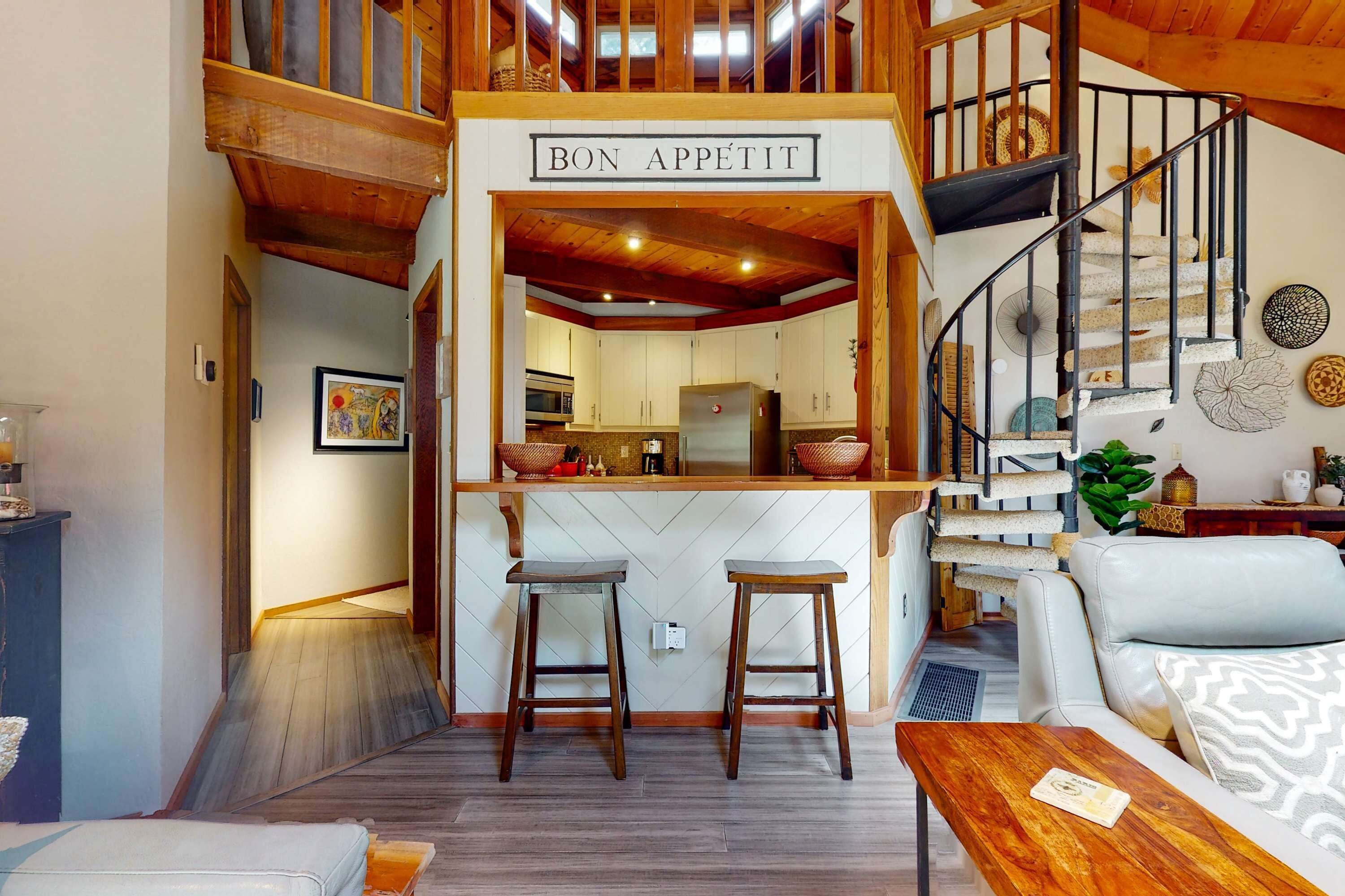 Un intérieur chaleureux avec un escalier en bois, un coin cuisine et un espace salon agrémenté d’une pancarte sur laquelle on peut lire « BON APPÉTIT ».