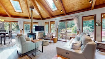 Un vaste salon doté d’un haut plafond, de poutres en bois, d’une cheminée et de grandes fenêtres offrant une vue sur l’extérieur.