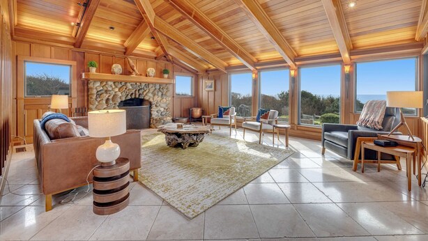 A spacious living area with a stone fireplace, wooden ceiling beams, and large windows offering a view of the outdoors. - Luxe Remodeled Oceanfront Home on the Bluff (Mendocino)