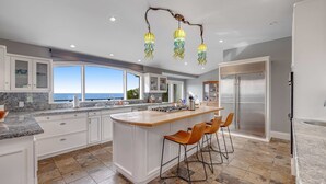 A modern kitchen with a large island, white cabinetry, and a view to the outdoors. - Luxe Remodeled Oceanfront Home on the Bluff (Mendocino)