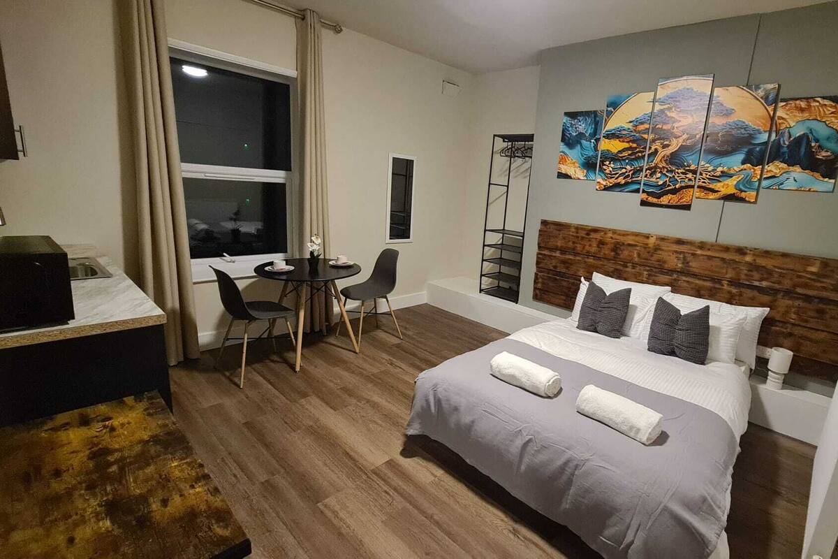 A hotel room with a bed, a desk, and chairs. There is a window with curtains, a painting on the wall, and a radiator.