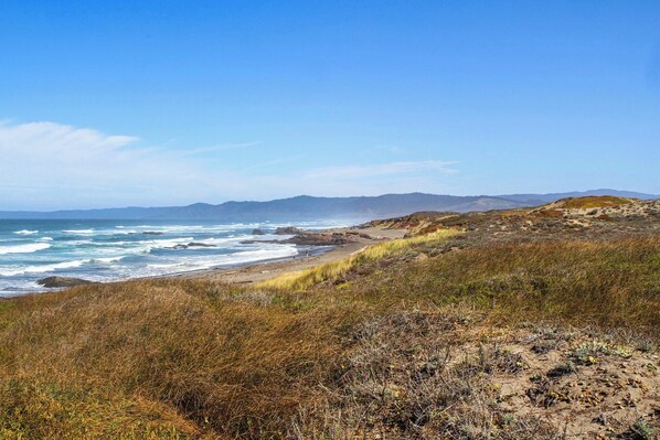 Paesaggio costiero con spiaggia, affioramenti rocciosi e montagne lontane sotto un cielo sereno.
