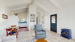 Amplia sala de estar con sofá, mesa de centro de madera y sillón azul. En el fondo, se distingue una zona de comedor con mesa y sillas. A la derecha, una puerta negra con ventanal, y en primer plano, una estufa azul.