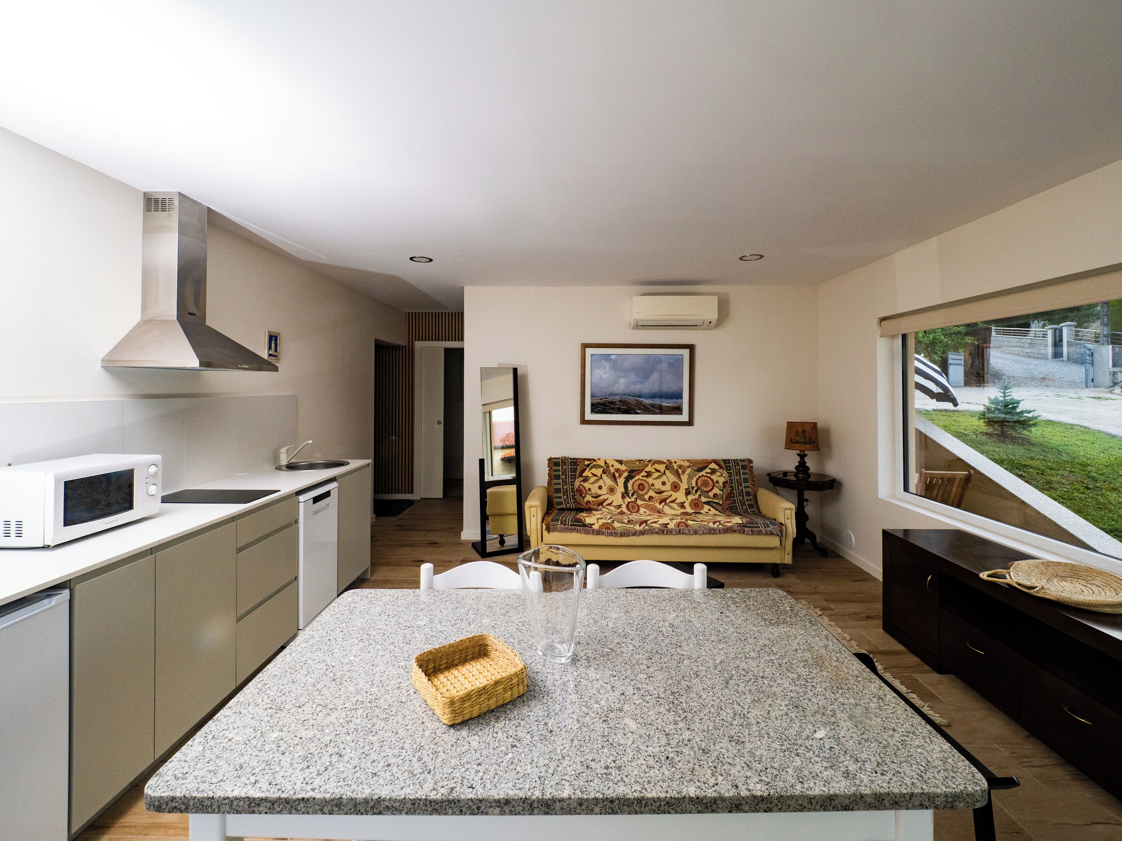 Cozinha compacta com micro-ondas, pia e exaustor de aço inoxidável. Uma pequena área de refeições com mesa e cadeiras de granito fica adjacente à cozinha. Na sala de estar, um sofá e uma luminária, com uma grande janela que oferece vista para o exterior.