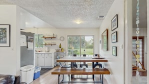 Una cocina moderna con armarios blancos, una mesa de comedor de madera y un banco. Hay cuadros en la pared y vistas al exterior a través de amplios ventanales.