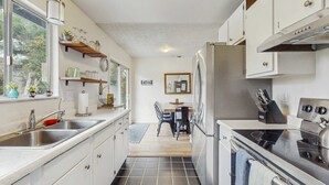 Una cocina compacta con armarios blancos, un refrigerador de acero inoxidable y una estufa negra. Se aprecia una ventana en el lado izquierdo, y un comedor con mesa y sillas al fondo.