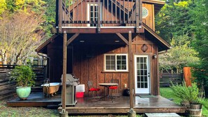 Una cabaña de madera con un balcón, una puerta principal y una pequeña zona exterior para sentarse con una mesa y sillas.