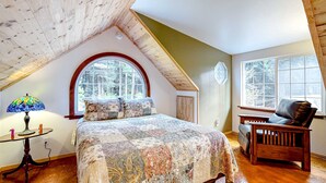 Un dormitorio con techo inclinado, una cama grande con colcha floral, una silla de madera, una mesa con lámpara y una ventana con vista a árboles.