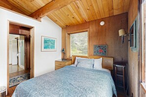Ein Schlafzimmer mit Holzboden, einem Bett mit gemusterten Betttextilien, einem Nachttisch, einem Fenster mit Jalousien und einer Wandlampe.