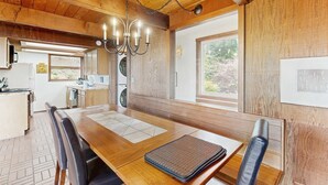 Une table à manger en bois, surmontée d’un lustre, avec une cuisine en arrière-plan et une fenêtre donnant sur des arbres.
