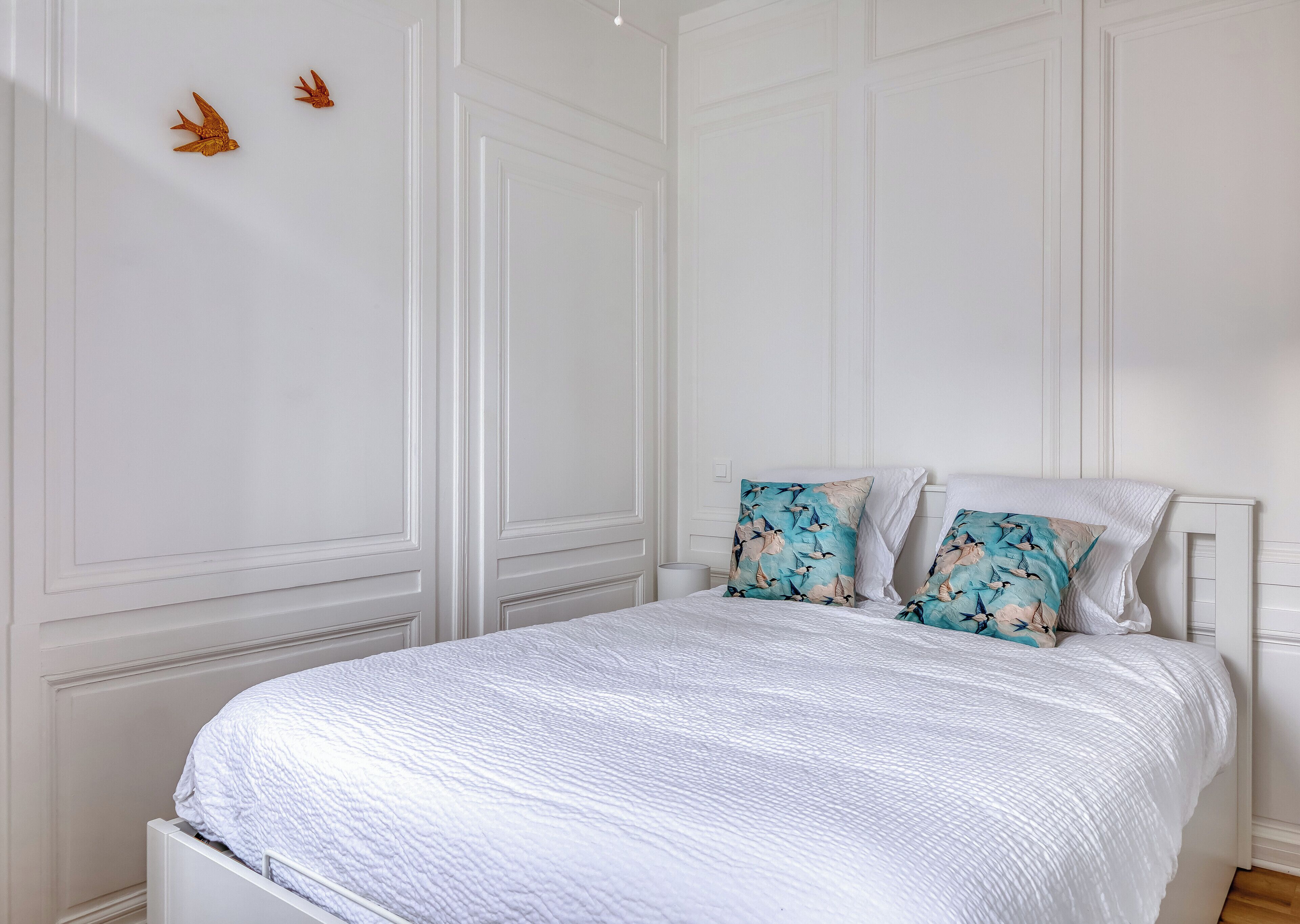 Ein Schlafzimmer mit einem weißen Bett, zwei Kissen mit Vogelmuster und einer weißen Tür mit dekorativen Verzierungen.