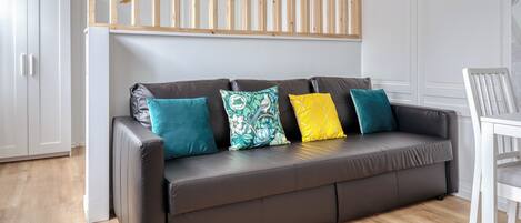 A modern living room with a dark brown leather sofa, four colorful cushions, a wooden slatted ceiling, and a white chair.