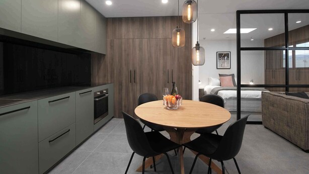 A modern kitchen with a dining area, featuring a round wooden table with black chairs, a fruit bowl, and pendant lights. - Treat yourself - luxury inner-city apartment (Dunedin)