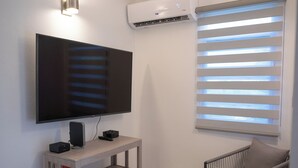Un salon avec un téléviseur à écran plat posé sur un meuble en bois, un climatiseur, une fenêtre avec des stores et un fauteuil.
