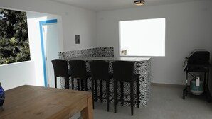 Une cuisine moderne dotée d’un îlot central, de tabourets de bar noirs, d’une table à manger en bois et d’une grande fenêtre qui laisse entrer la lumière naturelle.