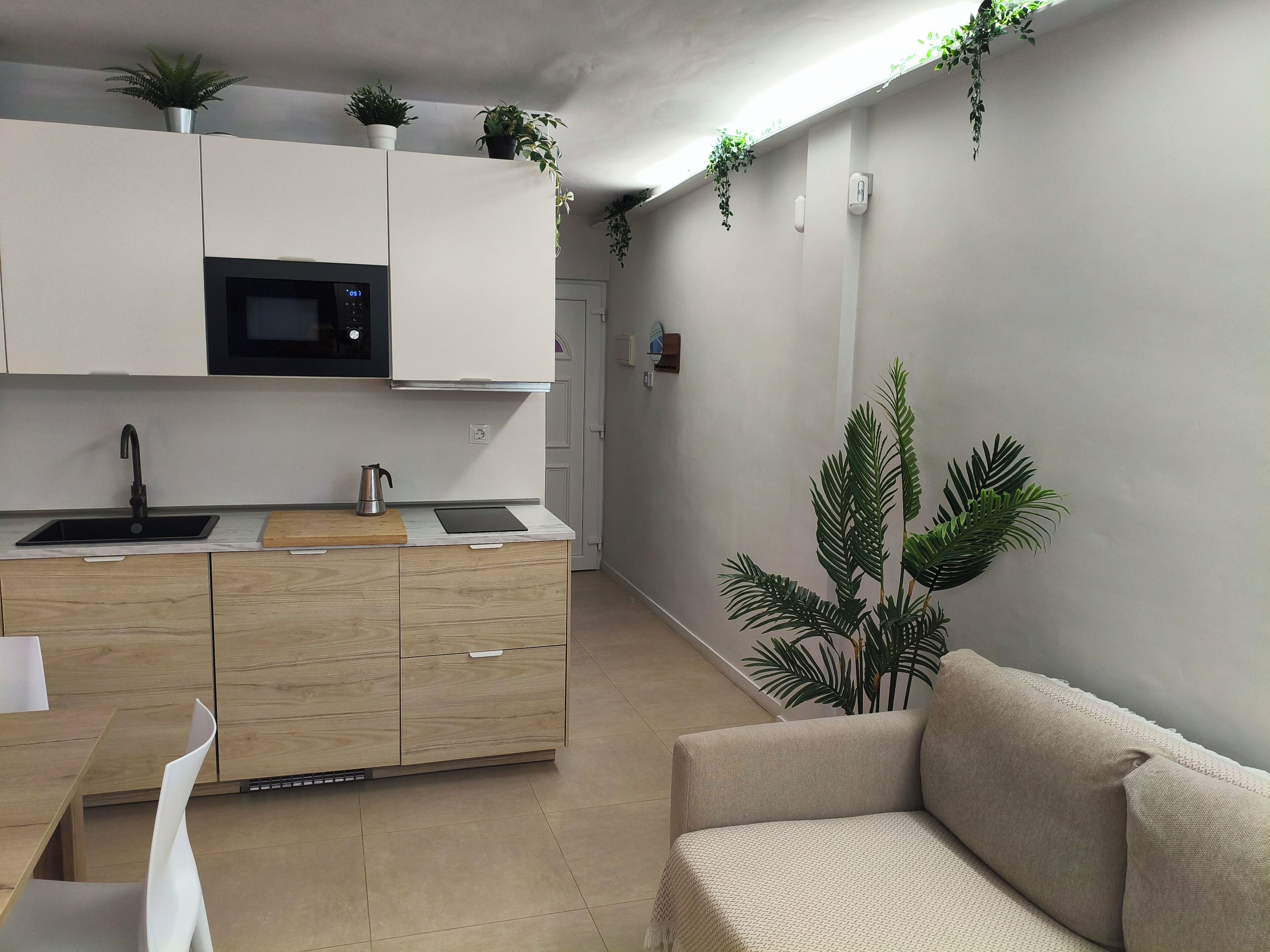 Eine moderne Küche mit weißen Schränken, einer schwarzen Mikrowelle und einem Spülbecken. Ein Holztisch mit einem weißen Stuhl steht im Essbereich. In der Wohnarea befindet sich ein beiges Sofa und rechts eine große Pflanze.