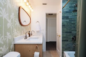 Ein Badezimmer mit einem weißen Waschbecken, einem Spiegel und einem Handtuch an der Wand. Es gibt einen Duschbereich mit Glastür und Handtuchhalter.
