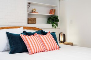 Ein ordentlich bezogenes Bett mit gestreiften und einfarbigen Kissen, eine Nachttischlampe, eine weiße Backsteinwand und ein Regal mit Büchern und einer Pflanze.