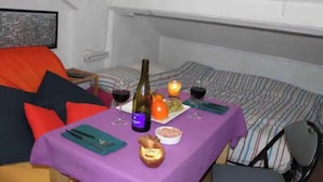 A small, cozy room with a bed, a table set for two with wine glasses, a bottle, and snacks. - Beau Studio Rue Didot (Paris)