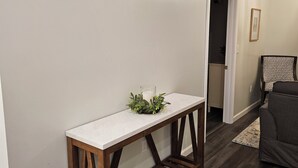 Un couloir avec une console en marbre blanc, un socle en lattes de bois et une petite composition florale. Un canapé en cuir marron foncé est visible au fond.