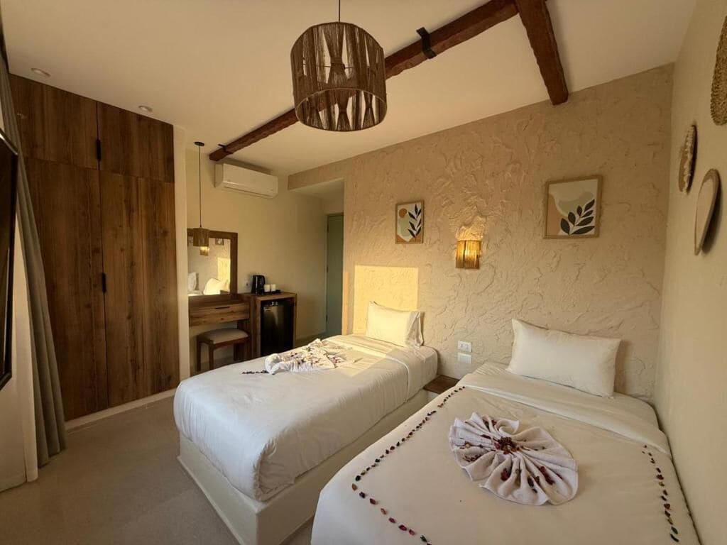 A hotel room with two beds, a wooden wardrobe, a desk with a chair, and a hanging light fixture.