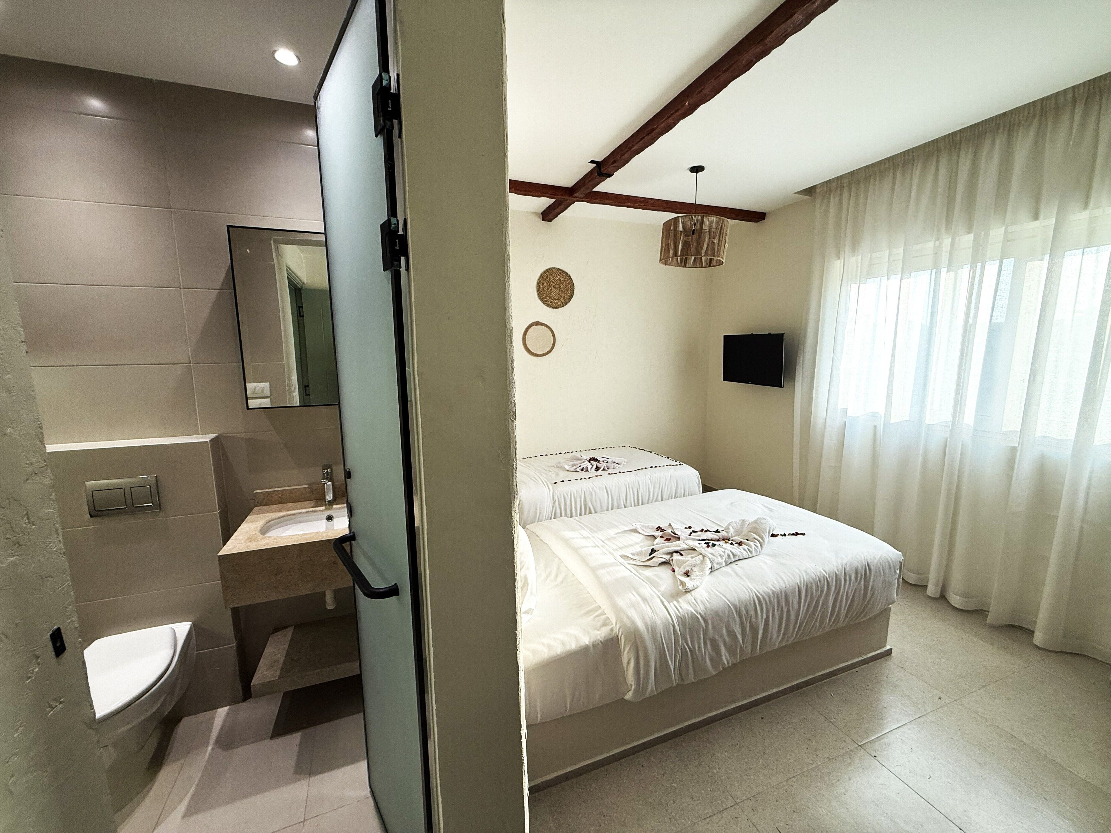 A modern hotel room with a bathroom, two beds, a TV, and a large window with curtains.