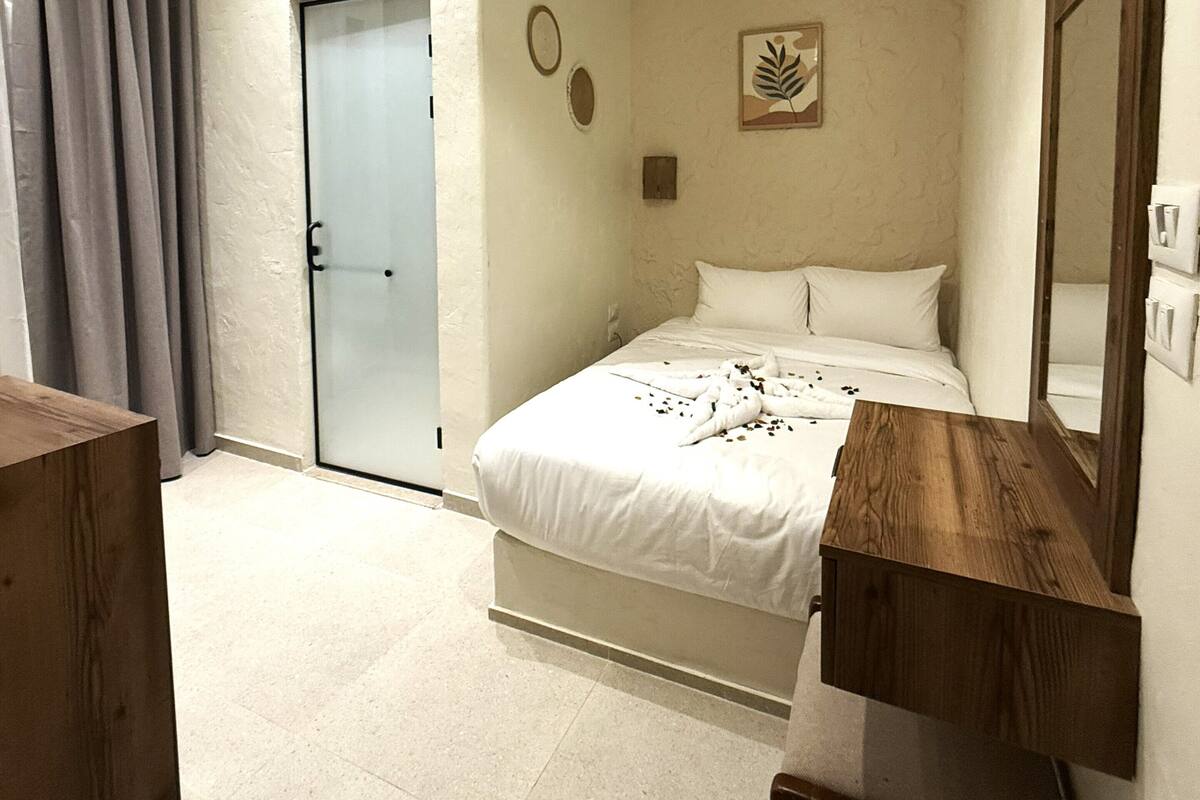 A bedroom with a bed, a wooden nightstand, a mirror, a ceiling light fixture, an air conditioning unit, and a door.