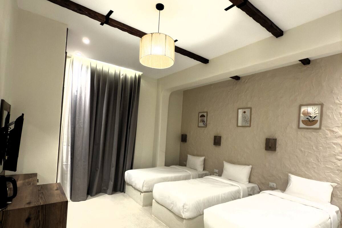 A hotel room with two beds, a wooden desk, a hanging light fixture, and a large window with curtains.
