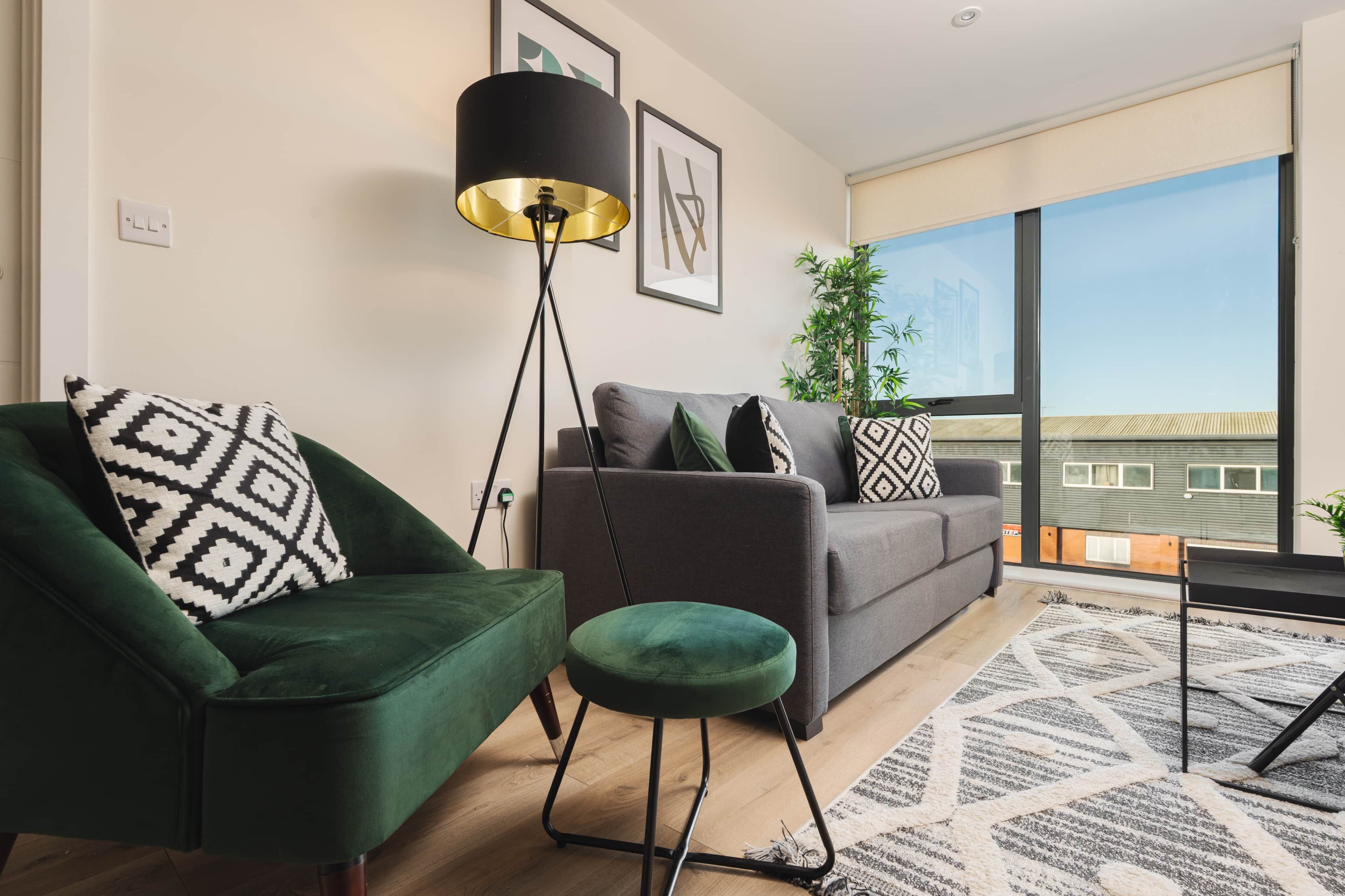 Ein modernes Wohnzimmer mit einem grünen Sessel, einem grauen Sofa, einer Stehlampe und einem großen Fenster mit Blick auf Gebäude.