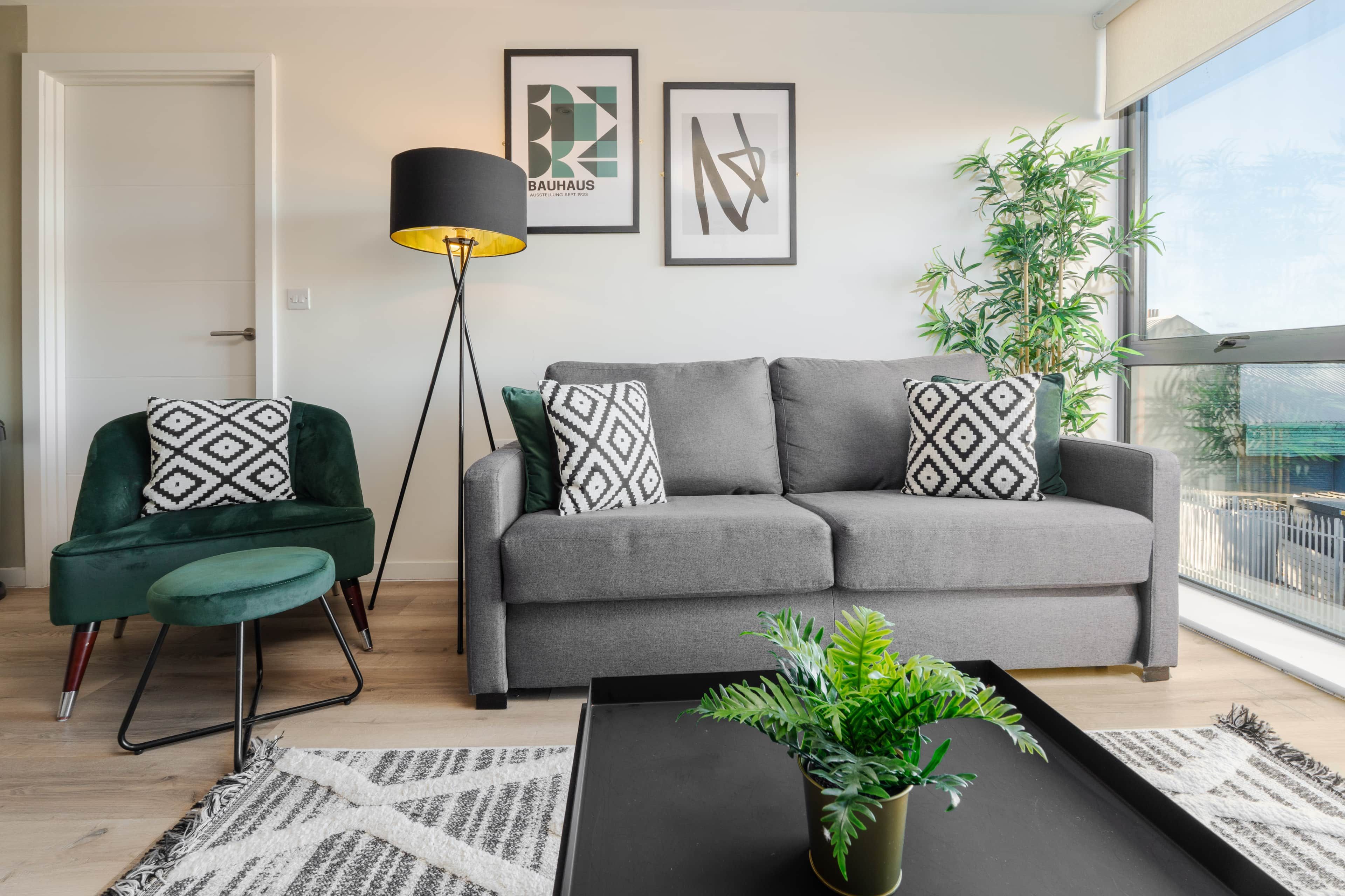A modern living room with a grey sofa, green armchair, and patterned cushions. There are two framed artworks on the wall, a floor lamp, and a potted plant on a black table.