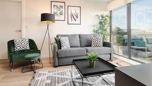 Ett modernt vardagsrum med en grå soffa, en grön fåtölj, ett svart soffbord och en golvlampa. På väggen hänger två inramade, abstrakta konstverk, och det finns ett stort fönster med utsikt över omgivningen.