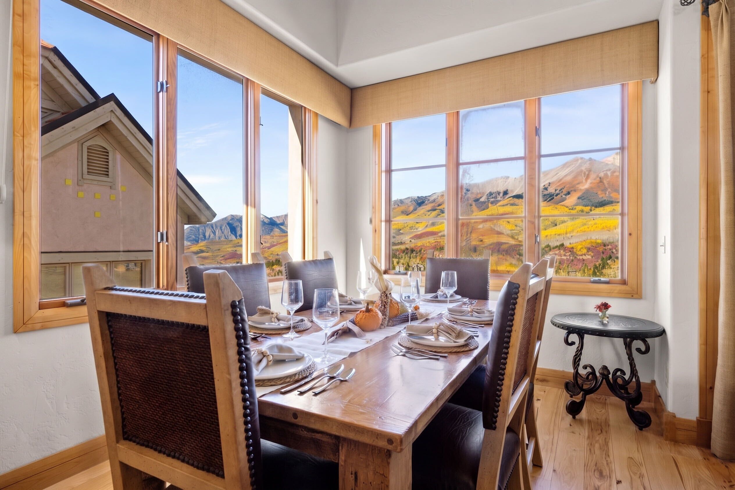 A dining area with a table set for two, large windows with a mountain view, and a small round table with a vase.