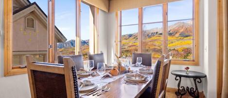 A dining area with a table set for two, large windows with a mountain view, and a small round table with a vase.