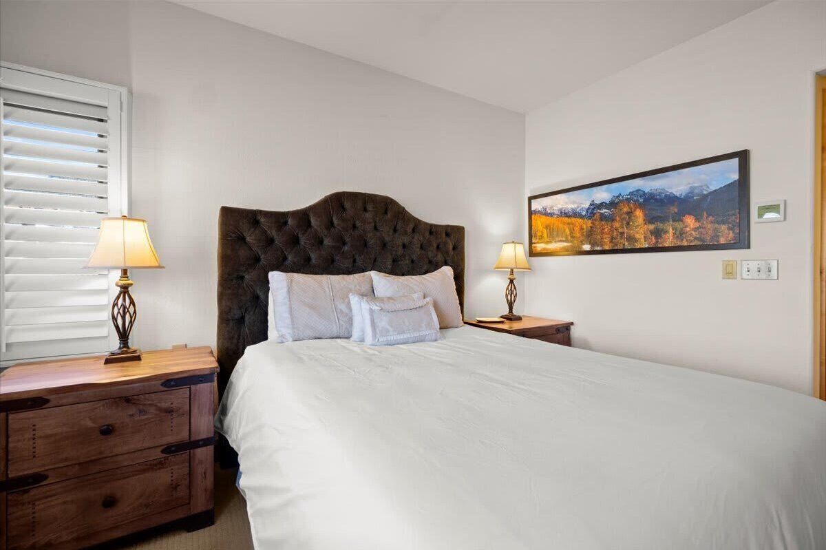 Un lit bien fait, doté d’une tête de lit capitonnée, deux tables de chevet avec des lampes et un tableau représentant un paysage, encadré et accroché au mur.