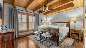 Ein Schlafzimmer mit Holzbalkendecke, einem großen Bett, einem Nachttisch, einem Fenster mit Jalousien und einem Teppich.