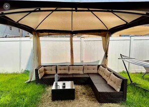 A covered outdoor seating area with a cushioned bench and a table, surrounded by a white fence.